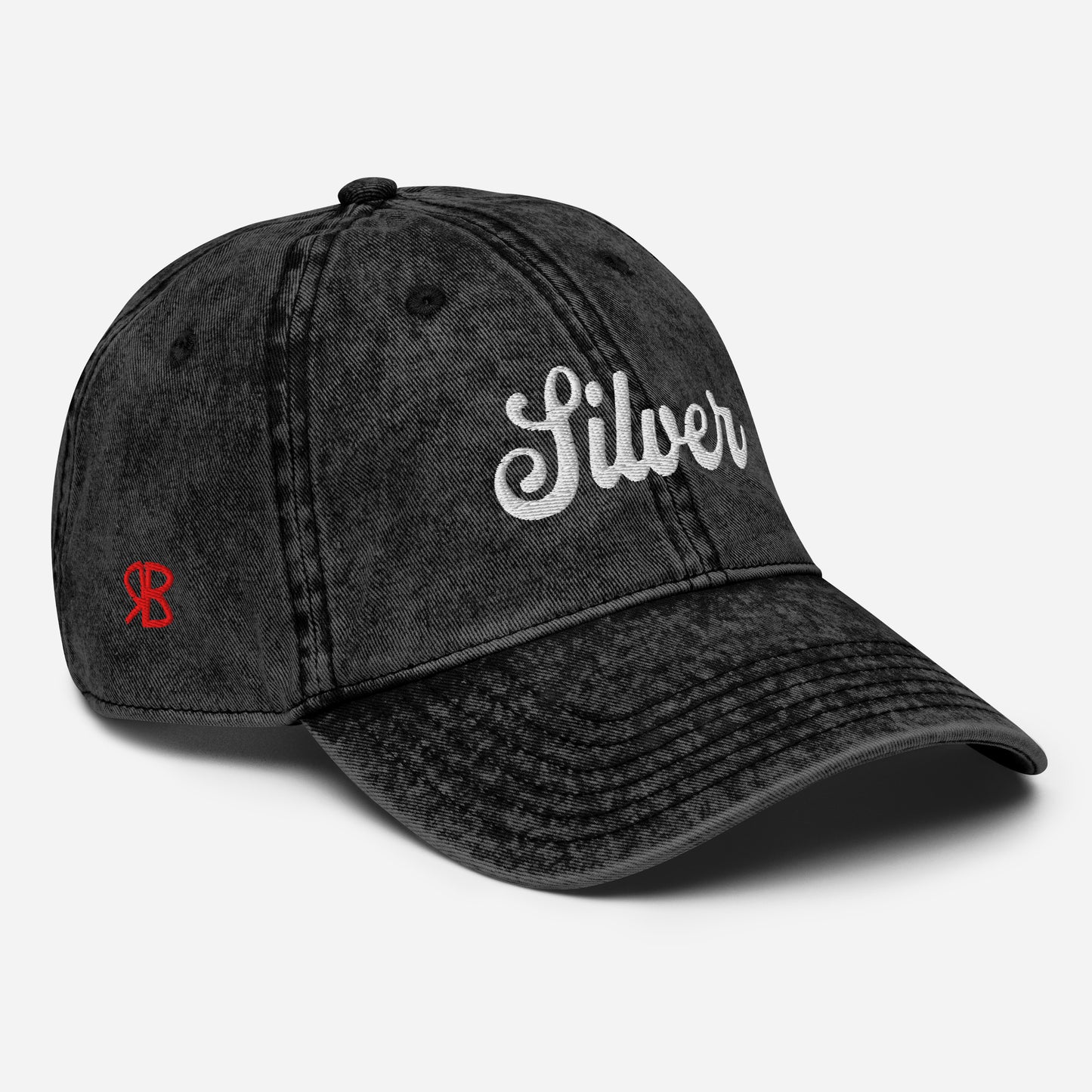 Flatlay 3/4 view of black denim vintage wash effect baseball cap with the word 'Raven' embroidered on the front in white adventure retro font with the initials RB in red which stand for the brand RedBlonde