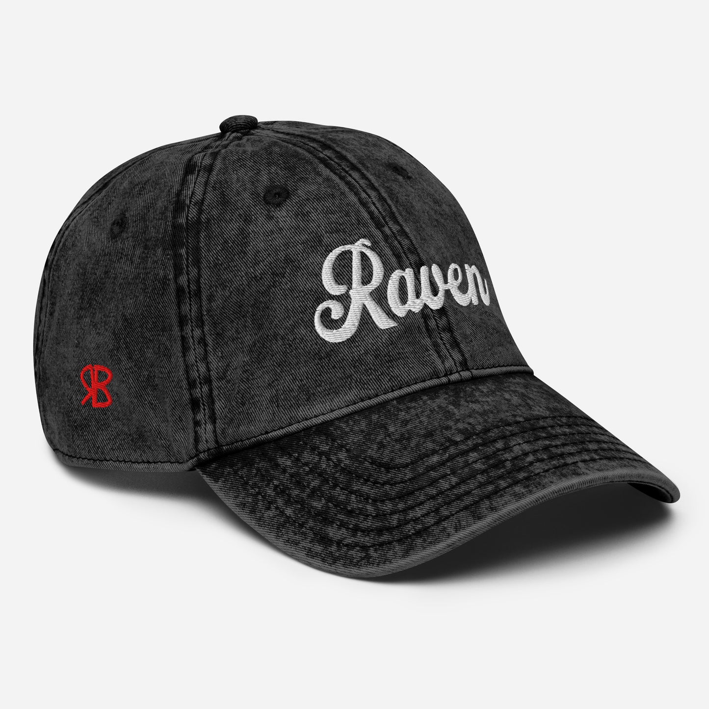 Flatlay 3/4 view of black denim vintage wash effect baseball cap with the word 'Raven' embroidered on the front in white adventure retro font with the initials RB in red which stand for the brand RedBlonde