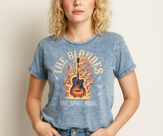 A blue denim wash unisex t-shirt with a graphic of a wooden guitar on fire with ‘THE BLONDES’ in Sancreek font above the burning guitar and ‘FIRE, ROCK & ROLL' underneath. Branded RedBlonde London in inside neck and right sleeve.