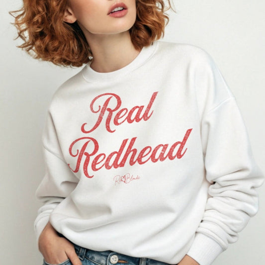 A redhead wears a classic white relaxed fit unisex sweatshirt with the words 'Real Redhead' written in red vintage milkstore font, like coco cola script. Branded RedBlonde London in red on inside neck and right sleeve
