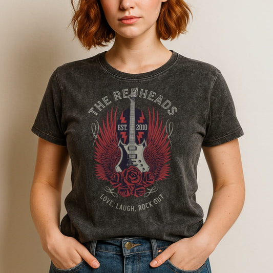 A redheaded woman wears a black denim wash unisex t-shirt with a graphic of a black & white guitar with red bird wings emerging from the side. The graphic features ‘THE REDHEADS' in Brookfield font in a semicircle above the guitar, while ‘Love, laugh, rock out’ is underneath. Branded RedBlonde London in inside neck and right sleeve
