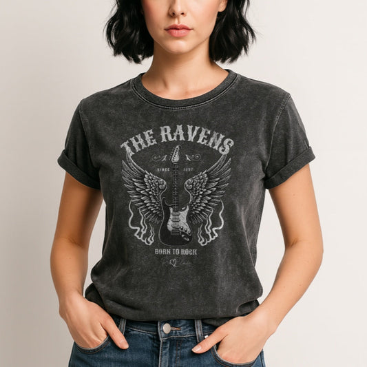 A woman with black hair wears a black denim wash unisex t-shirt with a graphic of a black & white guitar with raven bird wings emerging from the side. The graphic features ‘THE RAVENS’ in Sancreek font in a semicircle above the guitar, while ‘BORN TO ROCK & ROLL’ is underneath. Branded RedBlonde London in inside neck and right sleeve