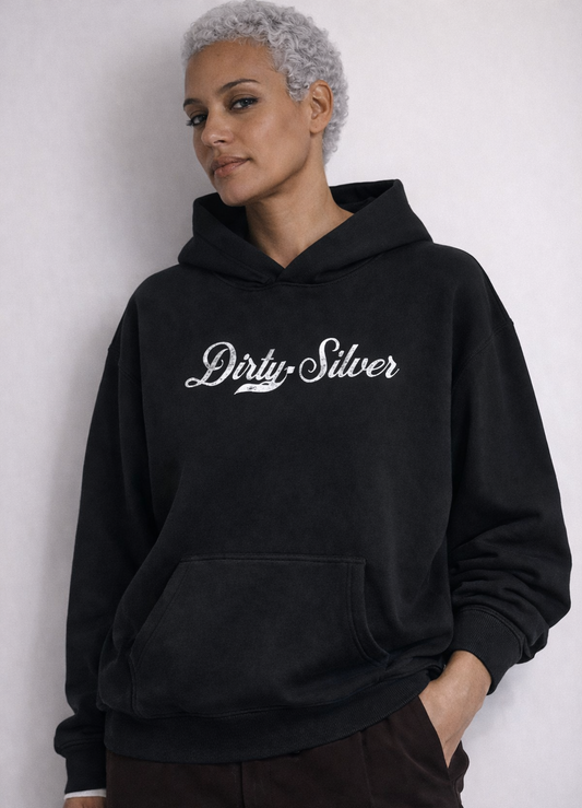 model wears a black pullover hoodie laid flat on a white background. It has a kangaroo front pocket and ribbed cuffs and hem. Across the chest is white Milkstore 01 script text reading “Dirty Silver”