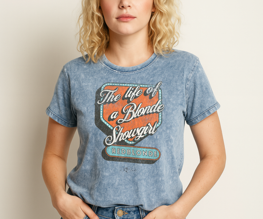 Blonde wears Vintage blue acid-wash unisex t-shirt with bold retro-style graphic text reading 'The life of a Blonde Showgirl' in white script over an orange and turquoise marquee design. The word 'RedBlonde' is displayed in turquoise block letters at the bottom, with subtle worn details giving the shirt a distressed, retro look." Branded redblonde on inside collar and right sleeve