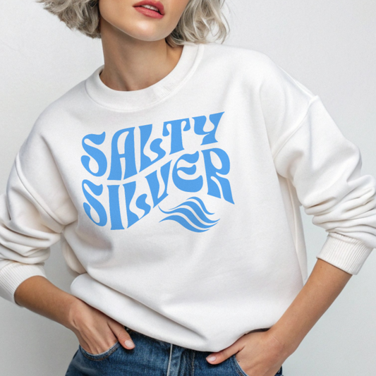Blonde wears a Classic relaxed fit unisex sweatshirt in white with 'Salty Silver' play on words slogan in blue retro 70s wavy text. Branded RedBlonde on inside neck and right cuff in red