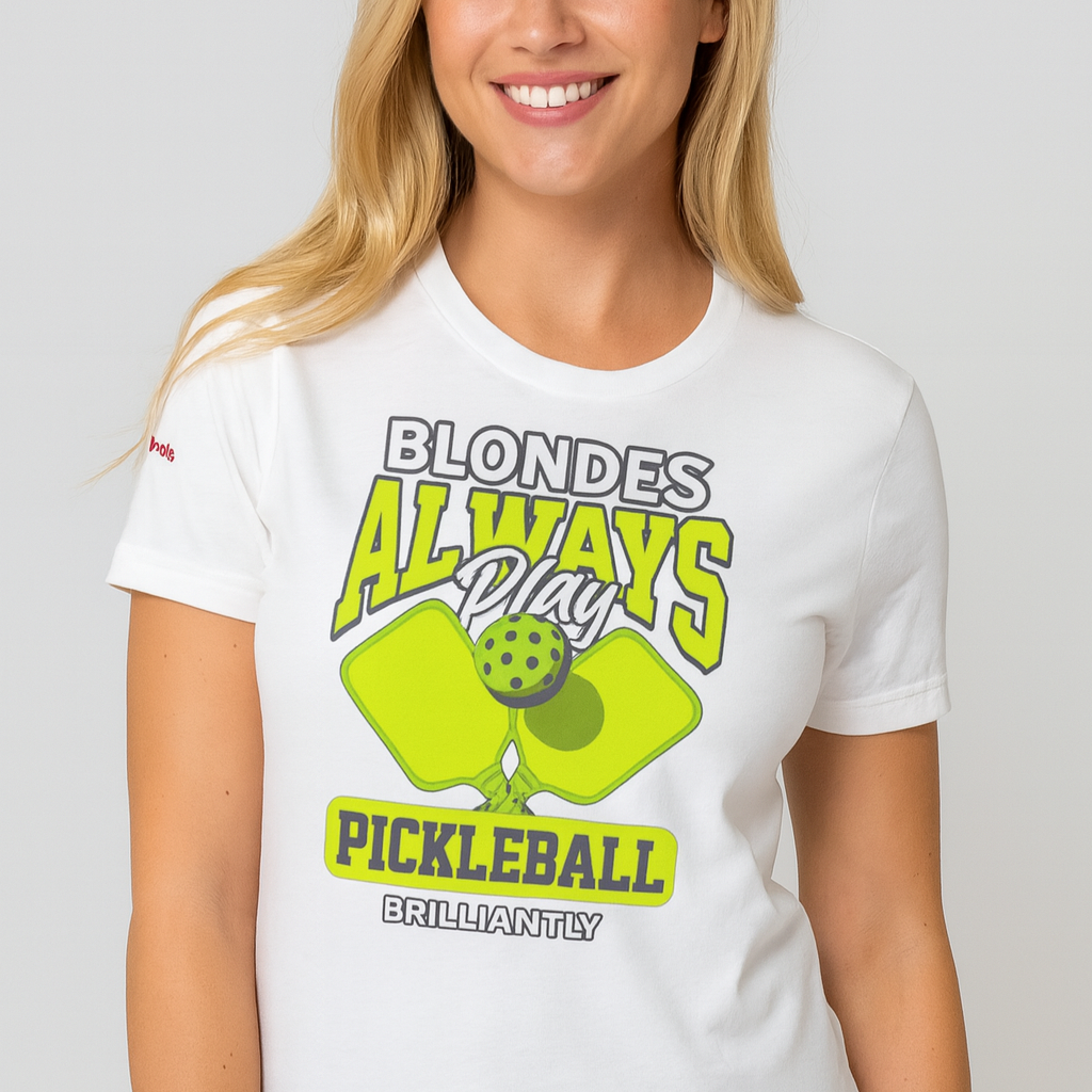Blonde wears a White unisex t-shirt featuring the phrase "Blondes Always Play Pickleball Brilliantly" in bold neon yellow and grey lettering, with two neon yellow pickleball paddles and a green pickleball in the center. Branded RedBlonde on inside the neck and on the right sleeve.