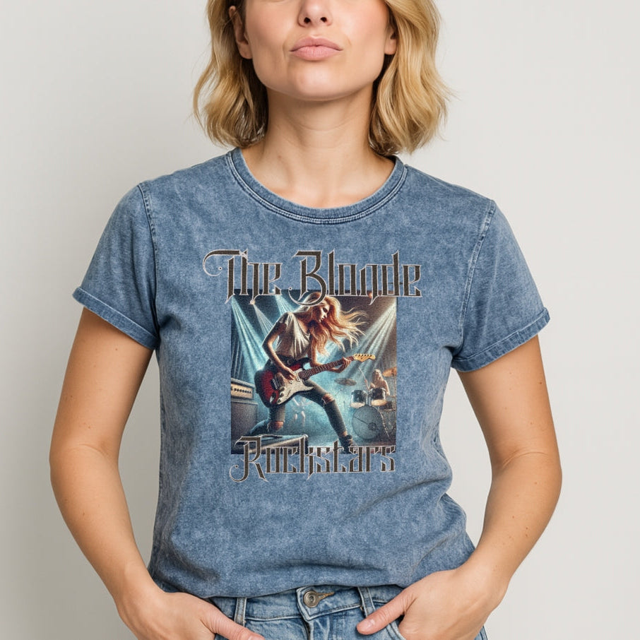 Blonde wears a blue denim wash unisex t-shirt with a graphic of a blonde female rock guitarist on stage with a drummer in the background, illuminated by stage lights. The text ‘The Blonde Rockstars’ is in bold, metallic avalen rekas font above and below the image. Branded RedBlonde London in red on the inside neck and right sleeve.