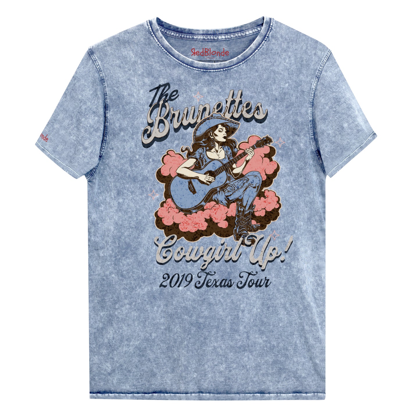Blue unisex t-shirt with a faded wash, featuring a retro graphic of a cowgirl playing a guitar surrounded by pink clouds. Text reads 'The Brunettes – Cowgirl Up! 2019 Texas Tour' in a Western-style font. Branded RedBlonde London on the inside neck and right sleeve