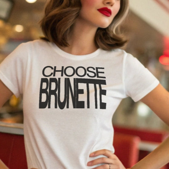 Brunette model leans on a white wall, wearing a 80s inspired white relaxed fit t-shirt with “Choose Brunette” in black helvetica now font emblazoned on the front.