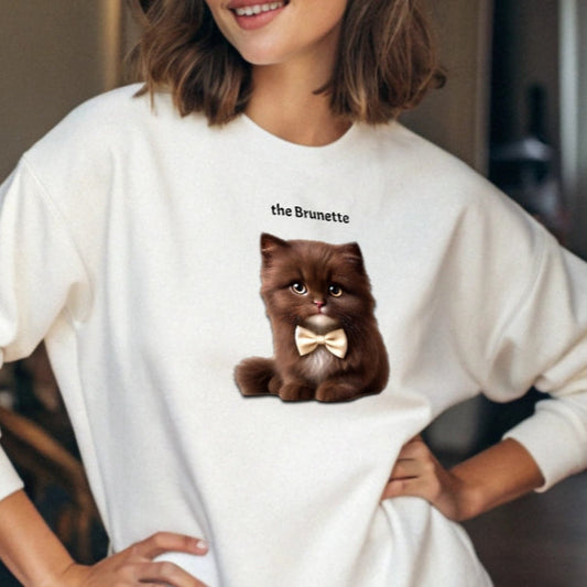 Brunette model wears white relaxed fit unisex sweatshirt with a cute graphic of a brunette kitten wearing a bow-tie and the words 'the Brunette' linking to Brunette hair. Branded RedBlonde in red on inside neck and right cuff