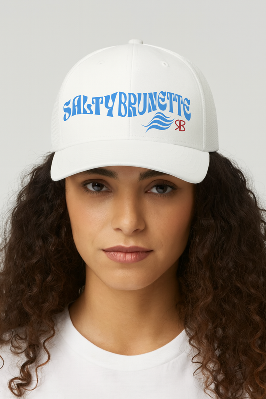 Brunette woman wears a white trucker cap with 'Salty Brunette' in blue Solino display font. Branded RB in red