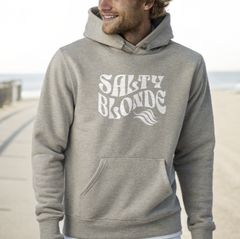blonde man on seafront wears a Classic relaxed fit unisex hoodie in athletic grey with 'Salty Blonde' in retro 70s text in white,
