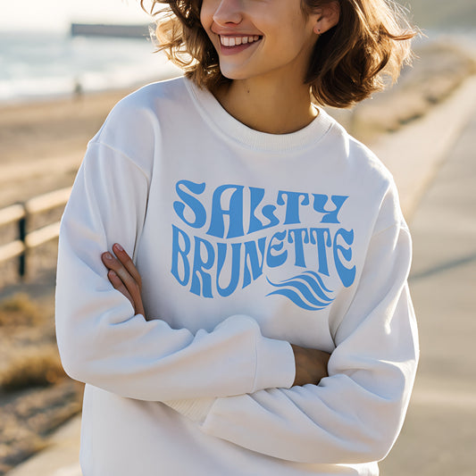 Classic relaxed fit unisex sweatshirt in white with 'Salty Brunette' play on words slogan in blue retro wavy 70s text. Worn by brunette woman on a seafront promenade