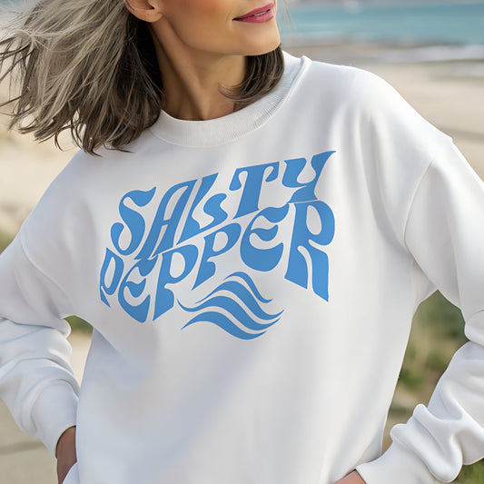 Classic relaxed fit unisex sweatshirt in white with 'Salty Pepper' play on words slogan in blue retro 70s wavy font. Worn by grey haired woman on a seafront promenade