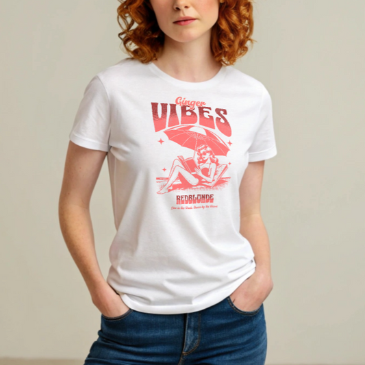 Ginger model wears a white unisex t-shirt featuring a retro-style red illustration of a redhead woman lounging under a beach umbrella reading a book, with bold text reading "Ginger Vibes" above and "RedBlonde" below, alongside the tagline "Live in the shade. Dance by the waves." Branded RedBlonde on inside neck and right sleeve