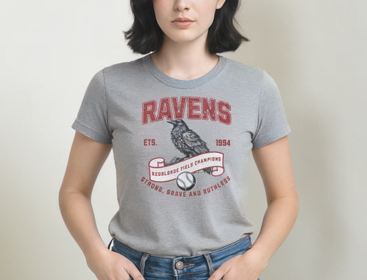 Light grey unisex t-shirt featuring a bold american collegiate vintage-style graphic with the word "RAVENS" in distressed red lettering. Below, a detailed black raven illustration perches on a scroll that reads "REDBLONDE FIELD CHAMPIONS," with a baseball beneath it. Additional text includes "ETS. 1994" and the phrase "STRONG, BRAVE AND RUTHLESS" in red. RedBlonde branding appears on the inner neck label and left sleeve in red text.
