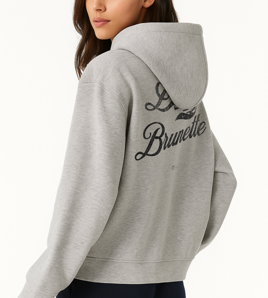 Model wears a grey hoodie with bold black Milkstore vintage-style script text “Dirty Brunette” across the back and a subtle RedBlonde signature logo underneath.