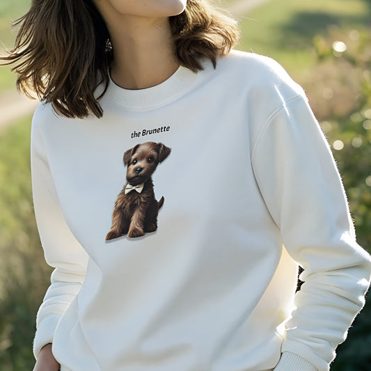 Model with brunette hair wearing white relaxed fit unisex sweatshirt with a cute graphic of a brunette puppy wearing a bow-tie and the words 'the Brunette' linking to brunette hair. Branded RedBlonde in red on inside neck and right cuff