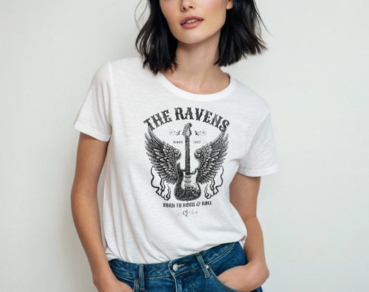 Raven haired woman wears a white unisex t-shirt with a graphic of a black & white guitar with raven bird wings emerging from the side. The graphic features ‘THE RAVENS’ in Sancreek font in a semicircle above the guitar, while ‘BORN TO ROCK & ROLL’ is underneath. Branded RedBlonde London in inside neck and right sleeve
