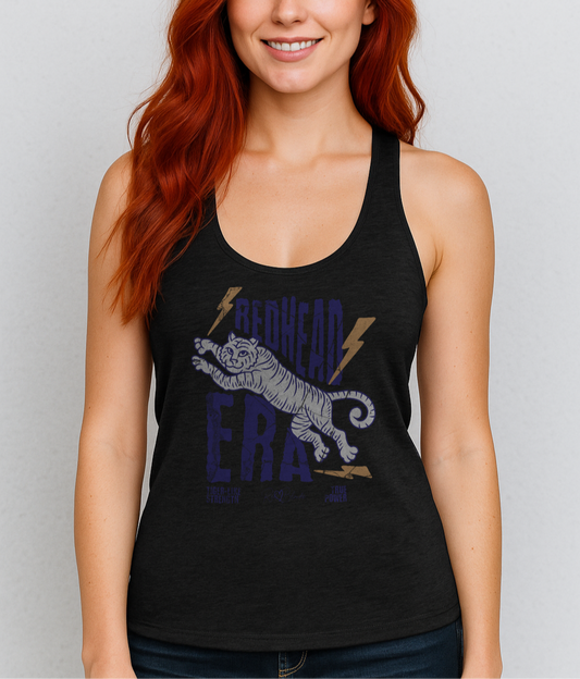 Redehead woman wears a vintage-style racerback tank in heathered black features a large, stylized illustration of a tiger leaping forward in pale purple and white. Bold, distressed text above and below reads “REDHEAD ERA” in uppercase purple letters, with two orange lightning bolts flanking the text. Beneath the lion in small purple letters are the phrases “Tiger-like strength” and “True power.” RedBlonde with a heart small signature-like logo appears below the text.