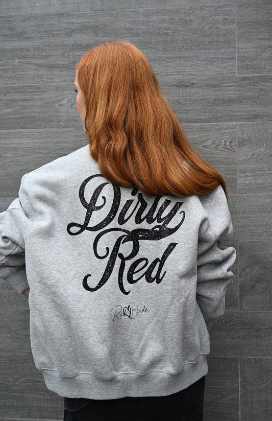 Back view of Redhead wears a Grey unisex zip up fleece bomber jacket featuring bold black milkstore script “Dirty Red” lettering and the RedBlonde logo printed on the back in a vintage script style.