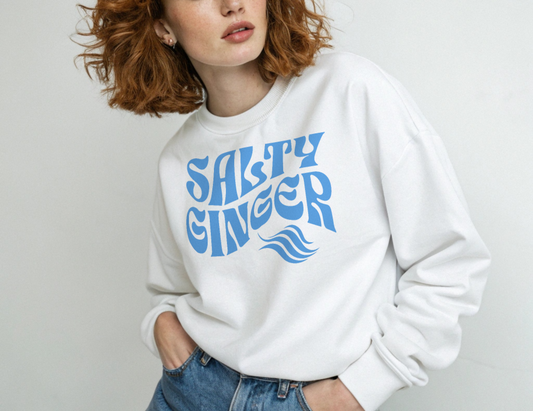 Redhead wears a Classic relaxed fit unisex sweatshirt in white with 'Salty Ginger' in blue Solino Display retro 70s wavy text. Branded RedBlonde London in red on inside neck and right cuff