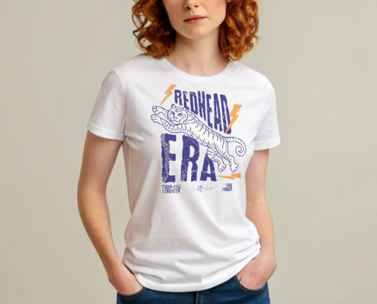 Redhead wears a White unisex short-sleeve t-shirt featuring a bold graphic of a leaping tiger outlined in purple, surrounded by orange lightning bolts. The text “REDHEAD ERA” appears in large distressed purple letters, with smaller text reading “TIGER-LIKE STRENGTH” and “TRUE POWER” underneath. Signed RedBlonde. The brand name “RedBlonde” is printed in red on the inside collar label and on the right sleeve near the cuff.