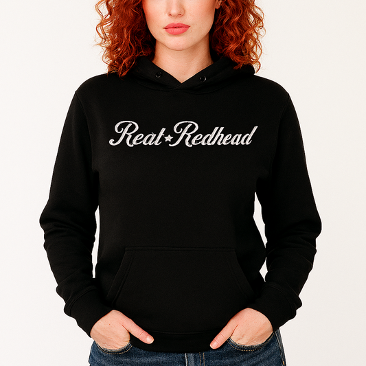 Redhead wears a classic black relaxed fit unisex hoodie with the words 'Real Redhead' written in white vintage milkstore font, like coco cola script. Branded RedBlonde London in red on inside neck and right sleeve