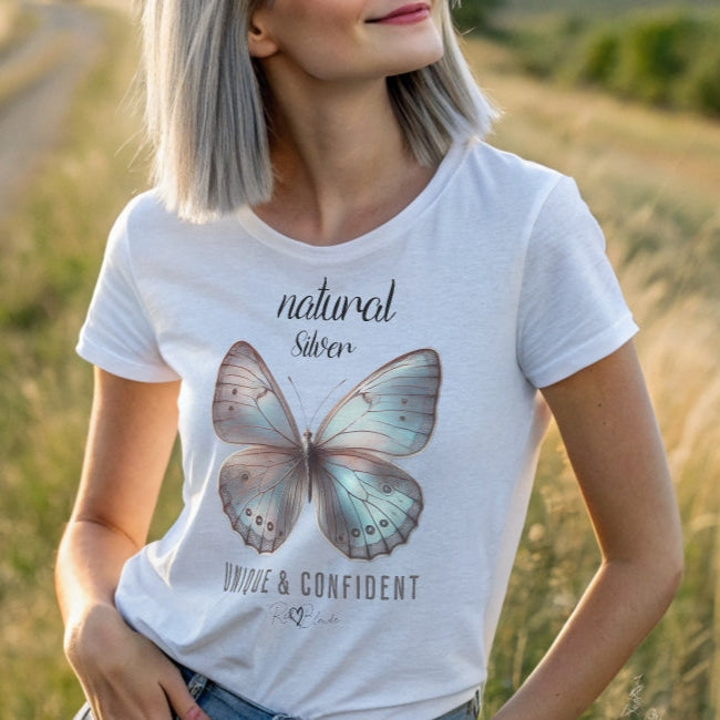 Silver gray haired woman wears white unisex relaxed fit t-shirt with the words natural Silver in arionna font above a graphic of a silver butterfly with 'UNIQUE & CONFIDENT' below in adventurer 06 font signed off by RedBlonde.