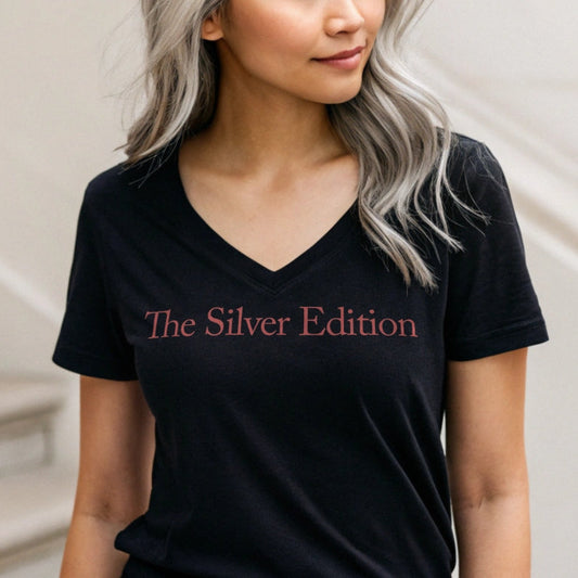 Silver / grey hair youthful woman wears a relaxed but slightly fitted black v-neck t-shirt with the words 'The Silver Edition' in red. Branded RedBlonde in red on the inside neck and right sleeve