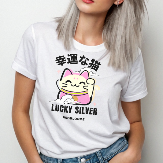 Silver haired model wears a white unisex T-shirt featuring a colourful illustration of a lucky cat (Maneki-neko) with pink ears and accents, smiling and waving one paw. Above the cat are Japanese characters reading "幸運な猫" (Lucky Cat), and below it are the words "LUCKY SILVER" in bold black font, followed by "REDBLONDE" in smaller text. The design is playful and cute, with stars and a cloud adding charm. The RedBlonde brand name appears in red on the inside label and on the right sleeve.