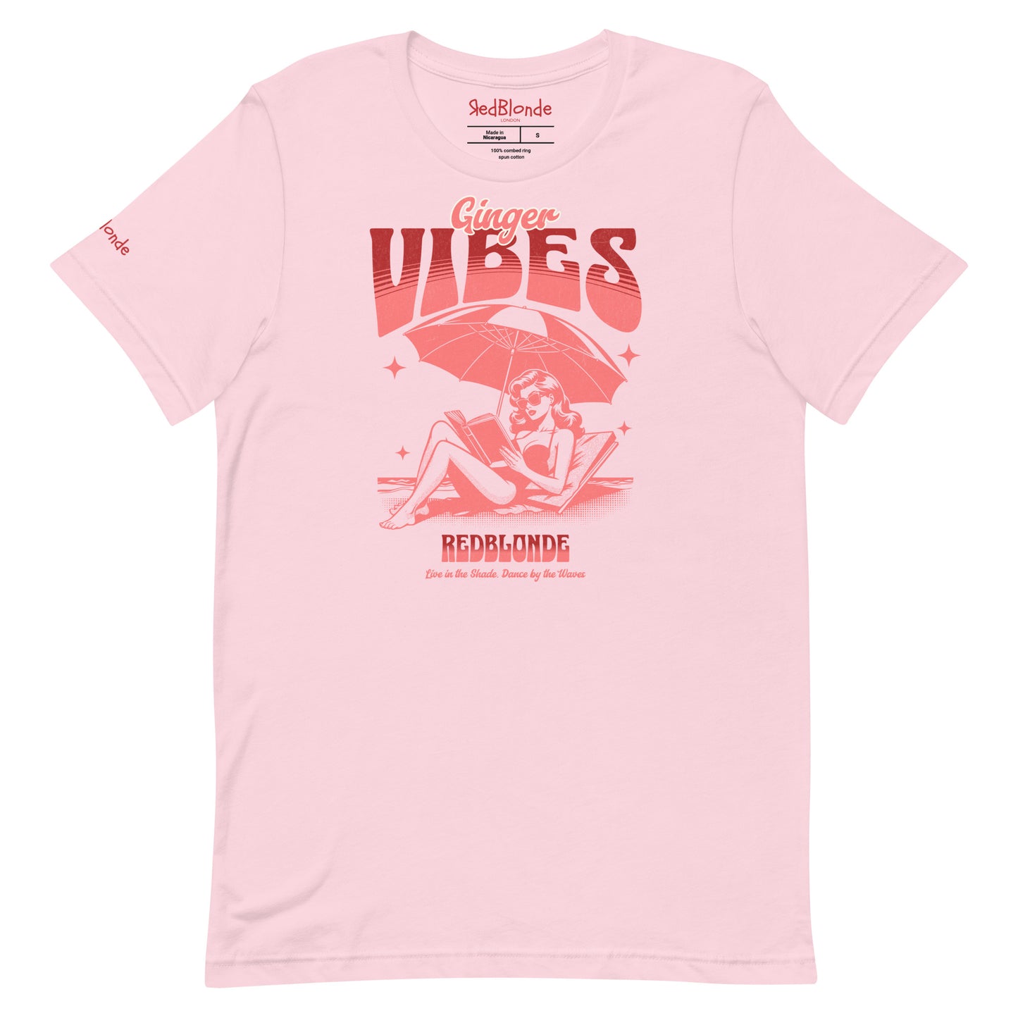Soft pink unisex t-shirt featuring a retro-style red illustration of a redhead woman lounging under a beach umbrella reading a book, with bold text reading "Ginger Vibes" above and "RedBlonde" below, alongside the tagline "Live in the shade. Dance by the waves." Branded RedBlonde on inside neck and right sleeve
