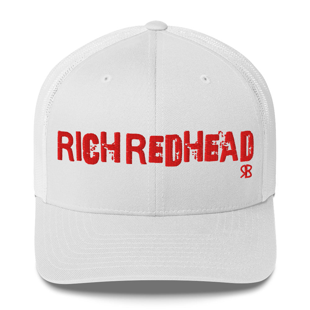 white mesh-back trucker cap with the bold distressed text “RICH REDHEAD” embroidered in red across the front, and a small red “RB” logo detail beneath the text. The cap has a structured front panel and curved brim