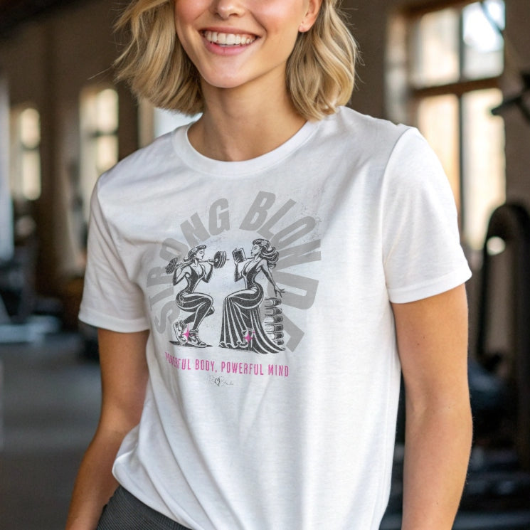 Woman wears a white relaxed-fit crew neck t-shirt featuring a graphic of two women: one holding a barbell on her shoulders, the other reading a book on a pile of books. The words ‘STRONG BLONDE’ are arced over the women in pale grey, while ‘Powerful Body, Powerful Mind’ is in bright pink underneath. The t-shirt is signed off RedBlonde with a heart. Branded RedBlonde in red on inside neck and right sleeve