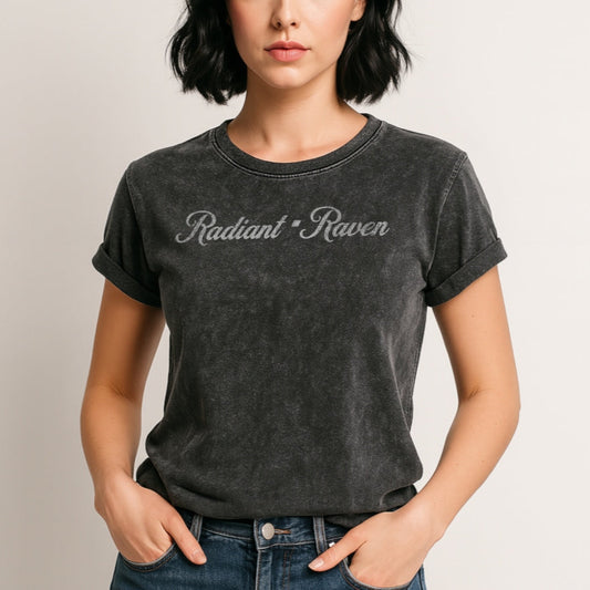 Woman with black hair wears a black denim wash vintage unisex relaxed fit slogan t-shirt with 'Radiant Raven' in milkshake script white font. Branded RedBlonde in red on inside neck and right sleeve