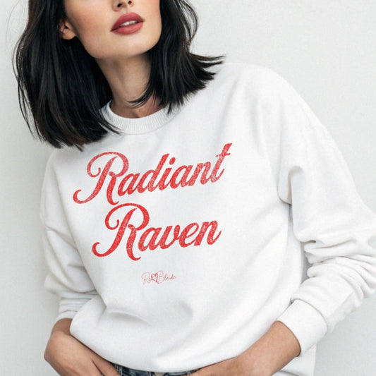 Women with jet black hair wears a classic white relaxed fit unisex sweatshirt with the words 'Radiant Raven' written in red vintage milkstore font, like coco cola script. Branded RedBlonde London in red on inside neck and right sleeve