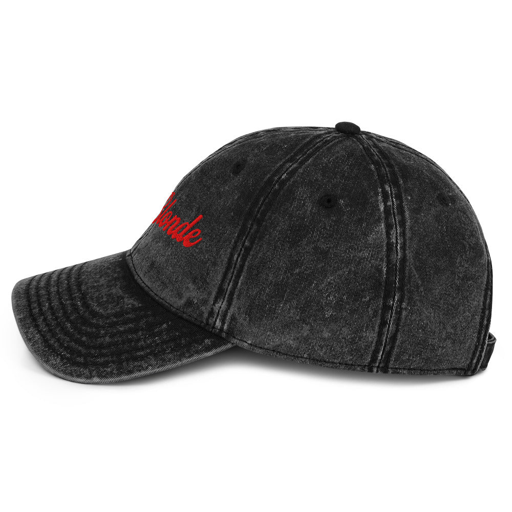 Flatlay side view of black denim vintage wash effect baseball cap with the word 'RedBlonde' embroidered on the front in red adventure retro font with the initials RB in red which stand for the brand RedBlonde