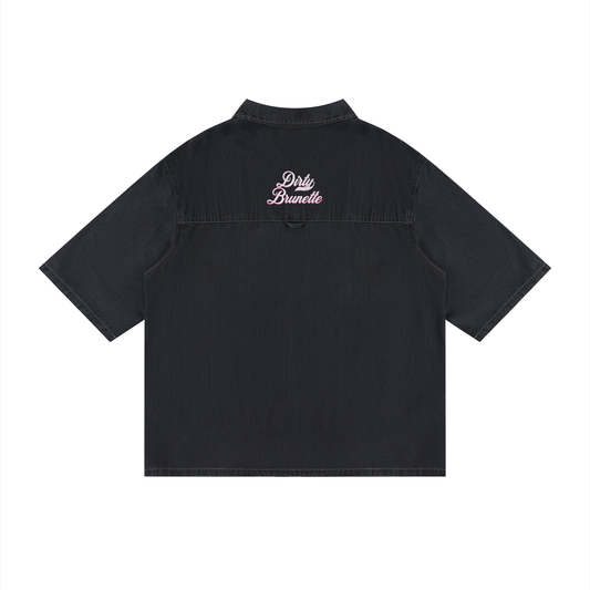 back view of a black, short-sleeve button-up shirt with a subtle faded finish and a small raised “Dirty Brunette” Milkstore script logo centered across the upper back.