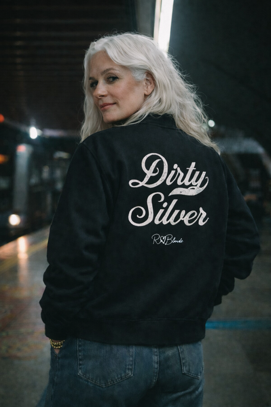 The back of a black crew neck zip up varsity jacket laid flat on a white background. It has ribbed cuffs, collar, and hem. Large white Milkstore 01 script text across the back reads “Dirty Silver,” with a small cursive RedBlonde logo beneath.