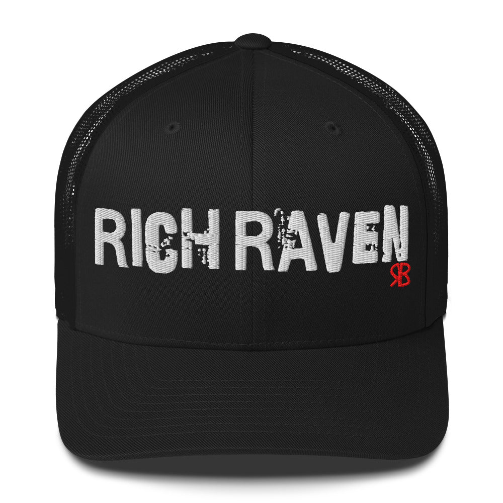 black mesh-back trucker cap with the bold distressed text “RICH RAVEN” embroidered in white across the front, and a small red “RB” logo detail beneath the text. The cap has a structured front panel and curved brim