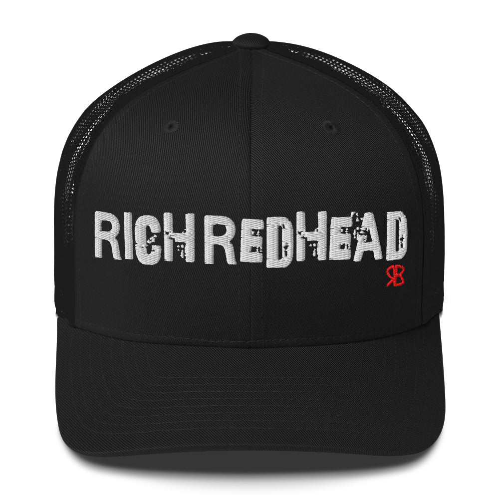 black trucker cap with the bold distressed text “RICH REDHEAD” embroidered in white across the front, and a small red “RB” logo detail beneath the text. The cap has a structured front panel and curved brim