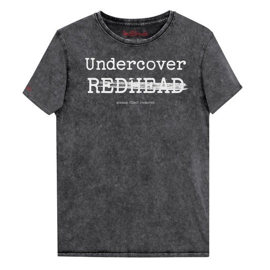 A black unisex denim-style t-shirt with a vintage washed finish. The front features bold white text reading “Undercover REDHEAD” partially struck through by a horizontal white bar. Beneath it, smaller text says “mission file // redacted” along with a subtle RedBlonde logo. The shirt has a classic crew neck and short sleeves with visible stitching for a rugged look. Branded RedBlonde London in red on inside neck and right sleeve