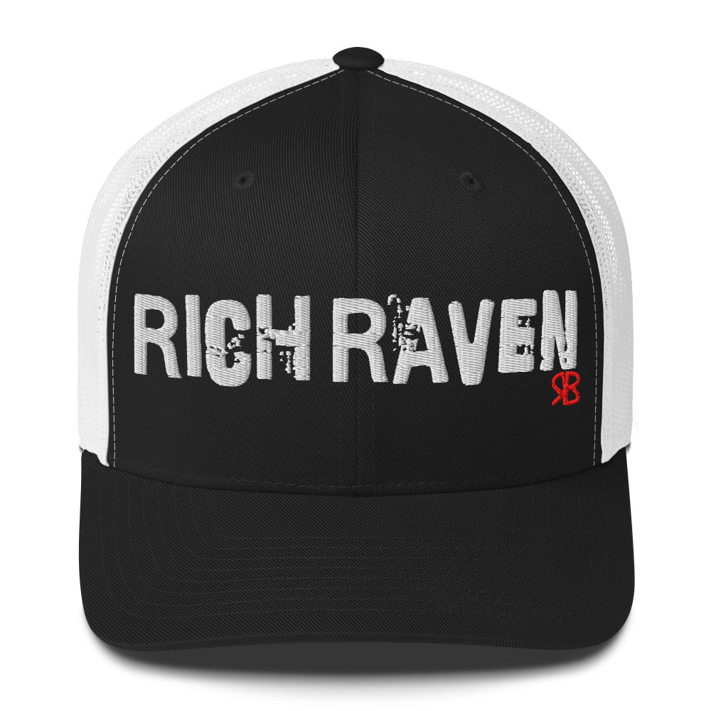 black/white mesh-back trucker cap with the bold distressed text “RICH RAVEN” embroidered in white across the front, and a small red “RB” logo detail beneath the text. The cap has a structured front panel and curved brim