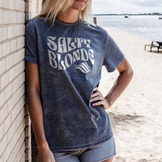 Blonde model, with her hand on her hip, leaning on a white wall on a beach with beach and sea in the background. She wears a soft denim vintage wash unisex graphic T-shirt with the slogan 'Salty Blonde' in white retro 70s wavy font