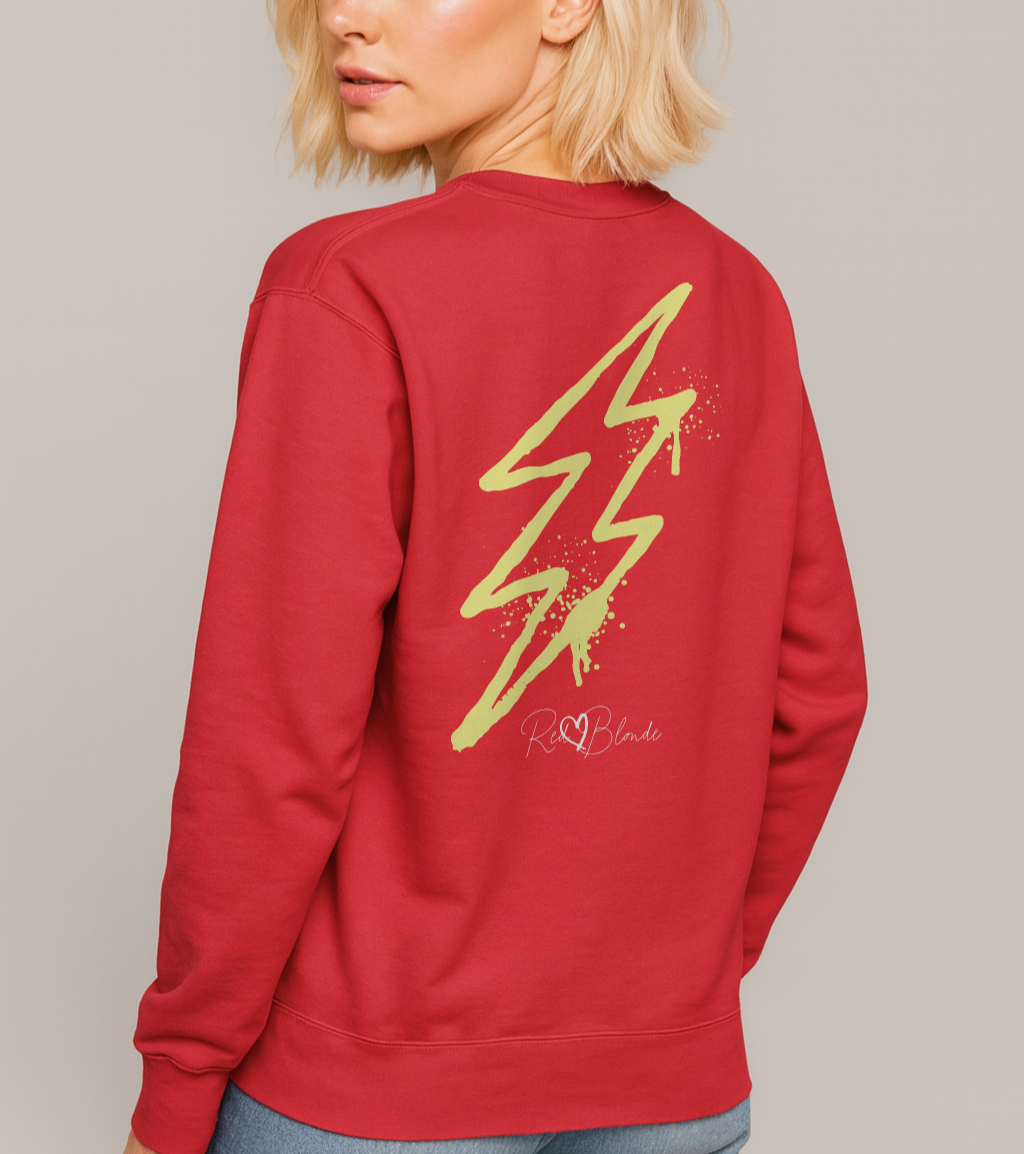 blonde wears a red unisex sweatshirt featuring a bold yellow lightning bolt graphic with paint-splatter detail and the RedBlonde logo printed below. The design has an edgy, electric streetwear vibe. Branded RedBlonde in red on right sleeve