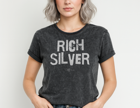 blonde wears a black acid-wash unisex denim T-shirt with a distressed vintage look. The shirt features the bold white text “RICH SILVER” in a weathered Ed Gein font, and features the RedBlonde signature logo. The shirt has short sleeves, a crew neckline, and visible seam detailing for a rugged, casual style. Branded Redblonde on inside neck and right sleeve