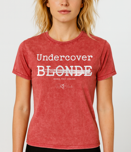 Blonde wears A red unisex denim-style t-shirt with a vintage washed finish. The front features bold white text reading “Undercover BLONDE” with “BLONDE” partially struck through by a horizontal white bar. Beneath it, smaller text says “mission file // redacted” along with a subtle RedBlonde logo. The shirt has a classic crew neck and short sleeves with visible stitching for a rugged look. Branded RedBlonde London in red on inside neck and right sleeve