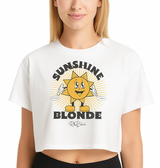 blonde woman wears a White women’s crop top with a retro cartoon-style yellow sun character, smiling with open arms, surrounded by radiant sunbeams. The text reads ‘Sunshine Blonde’ in bold vintage Belzebuth lettering, with small details including ‘ESTD 1994’ and the RedBlonde signature logo beneath."