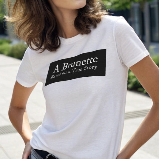 brunette model wears a white unisex relaxed fit t-shirt with the slogan 'A Brunette. Based on a True Story' in white adobe caslon font in a black box graphic. Branded RedBlonde London in red on inside neck and right sleeve