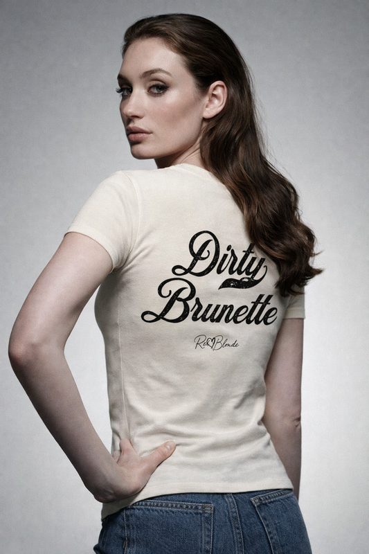 Brunette wears a fitted, short-sleeve off white T-shirt laid flat on a white background. It features large black Milkstore 01 script text reading “Dirty Brunette” across the upper back, with a small cursive “RedBlonde” logo beneath. The hem is slightly curved.